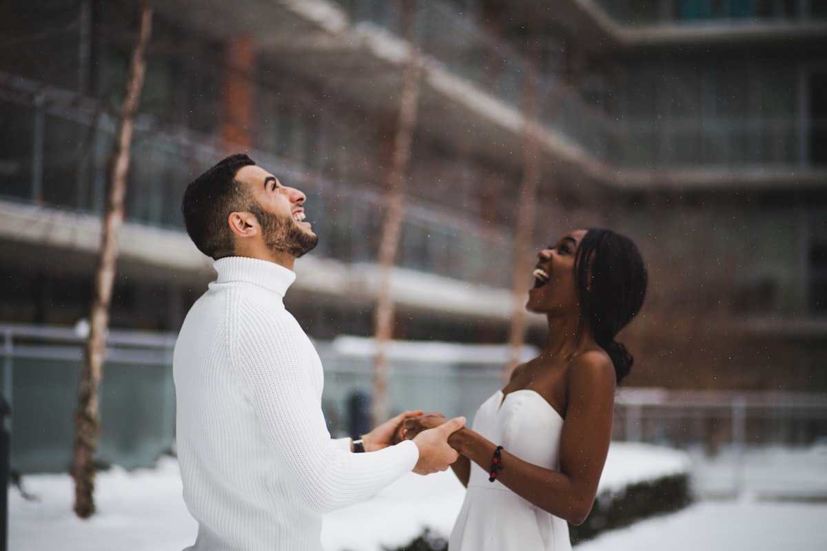 Happy diverse interracial couple holding hands and laughing together outdoors in the snow, the woman in a white dress