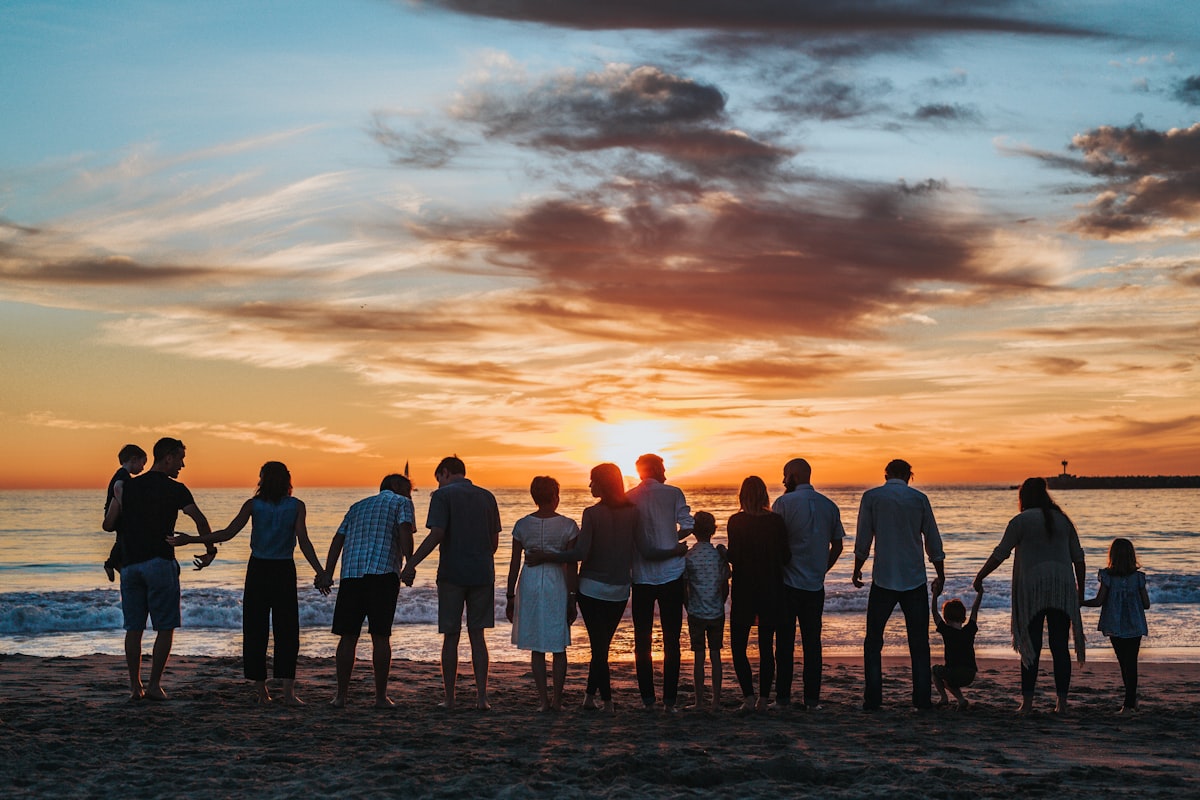 A large multigenerational family, from grandparents to young children, standing together and holding each other on a beach at warm golden sunset